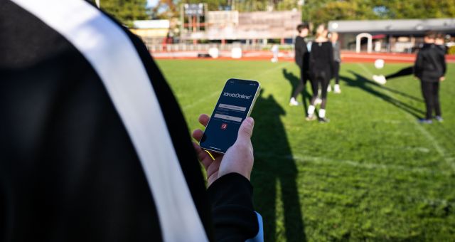 Person står med ryggen mot en fotbollsplan och kollar på mobilen